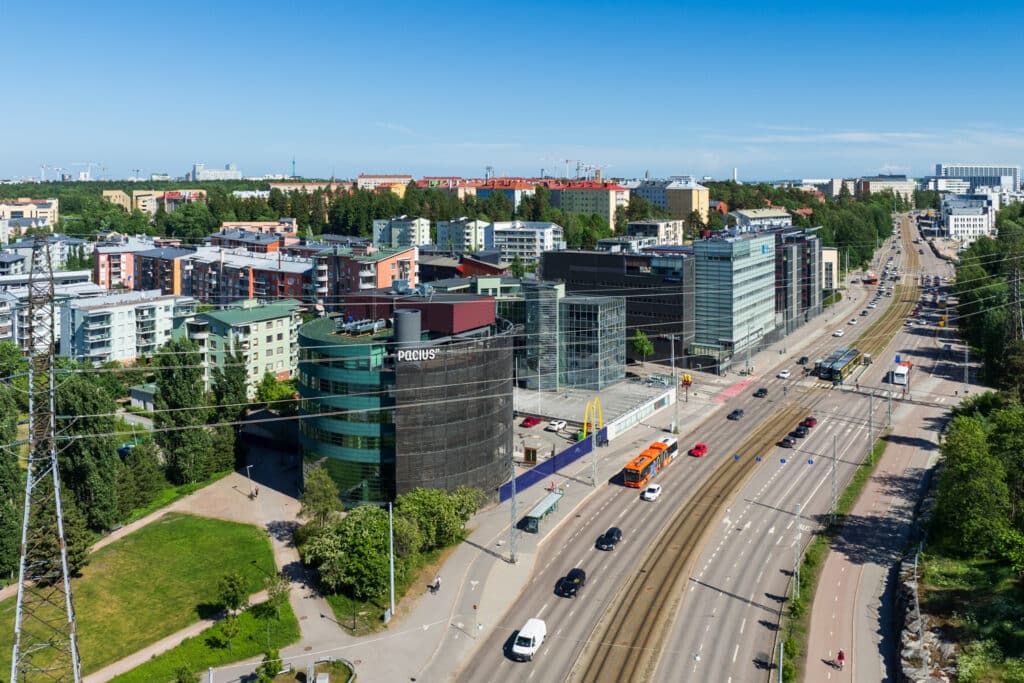 Helsingin Pikku-Huopalahden kerrostalokantaa ja vehreää puistoa ilmasta kuvattuna kesällä. Etualalla pyöreä Paciuksenkatu 29 toimitilatalo.
