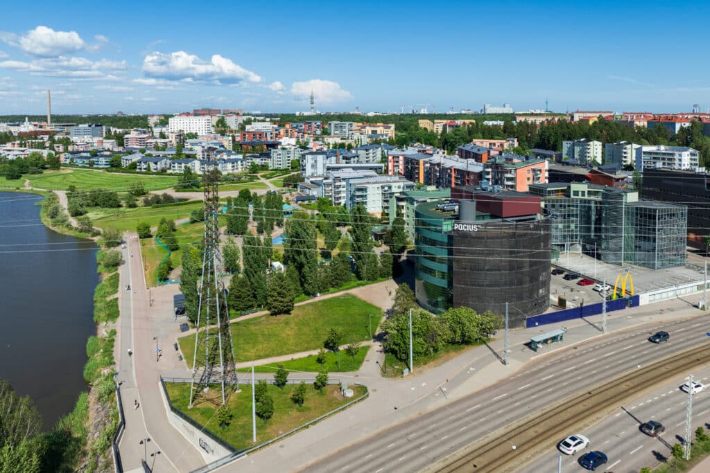 Helsingin Pikku-Huopalahden kerrostalokantaa ja vehreää puistoa ilmasta kuvattuna kesällä. Etualalla pyöreä Paciuksenkatu 29 toimitilatalo.